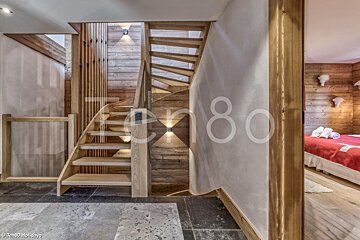 A wooden staircase in a rustic chalet with stone flooring, warm lighting, and a partial view of a bedroom on the right.