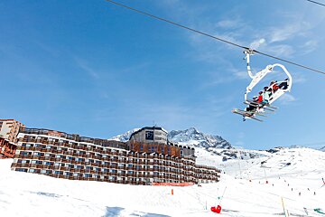 Skiers on a chairlift glide over a snowy mountain slope with a large resort building and snow-capped peaks under a clear blue sky.