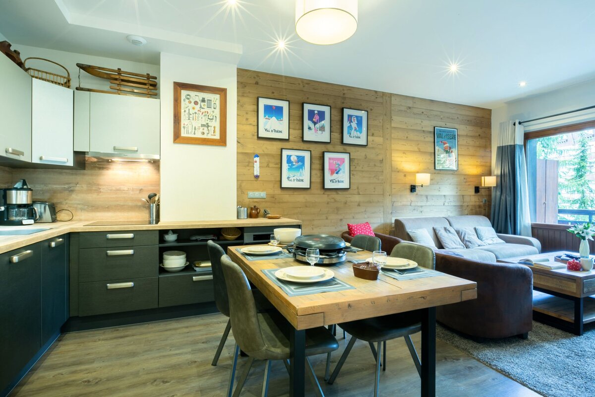 Modern open-plan kitchen, dining, and living space. Features a wooden accent wall with ski posters, a set dining table, and cozy sofas.
