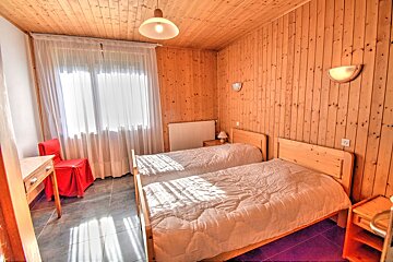 A wooden-paneled bedroom with two single beds, a bright window with white curtains, and a red chair. Natural light floods the space.