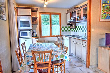 A cozy kitchen/dining area with a wooden table, patterned tablecloth, and wooden chairs. Features include various appliances, green & white tiled backsplash, and a window.