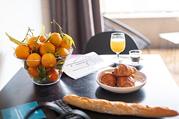 A bowl of oranges sits on a table next to croissants and a glass of orange juice