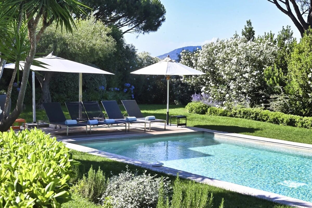 A sunny backyard swimming pool surrounded by lush green lawns, trees, sun loungers, and white patio umbrellas.