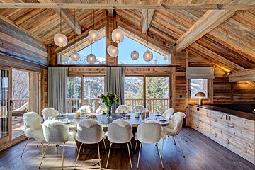 Luxurious, rustic wooden chalet dining room with fluffy chairs, a long table, and large windows showcasing snowy mountain views.