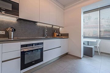 Modern white kitchen with dark backsplash, under-cabinet lighting, integrated appliances, and a compact dining area by a window with blinds.