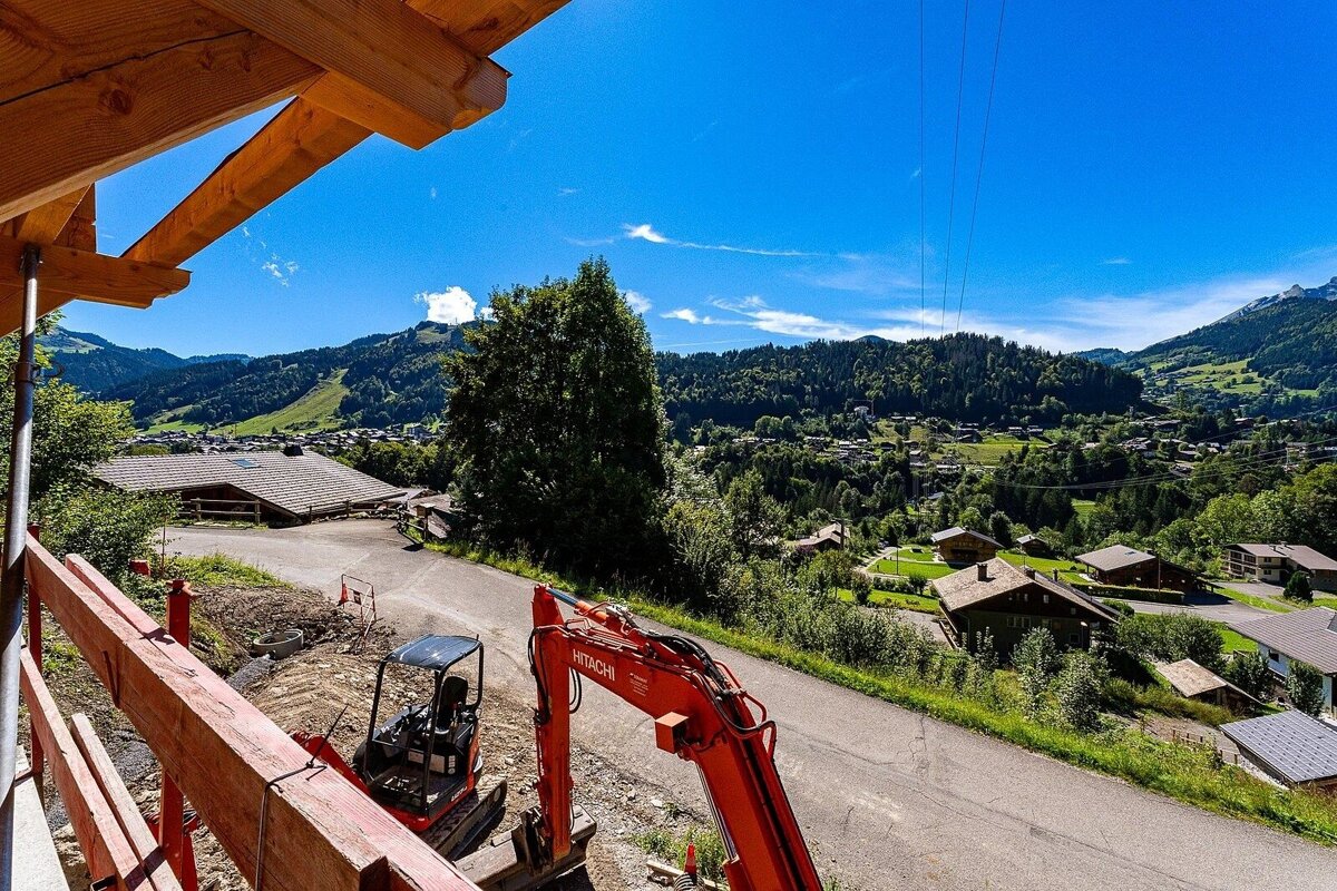 Vibrant mountain village scene with lush green hills and scattered houses under a clear blue sky. Construction elements, including excavators and wooden beams, are in the foreground.