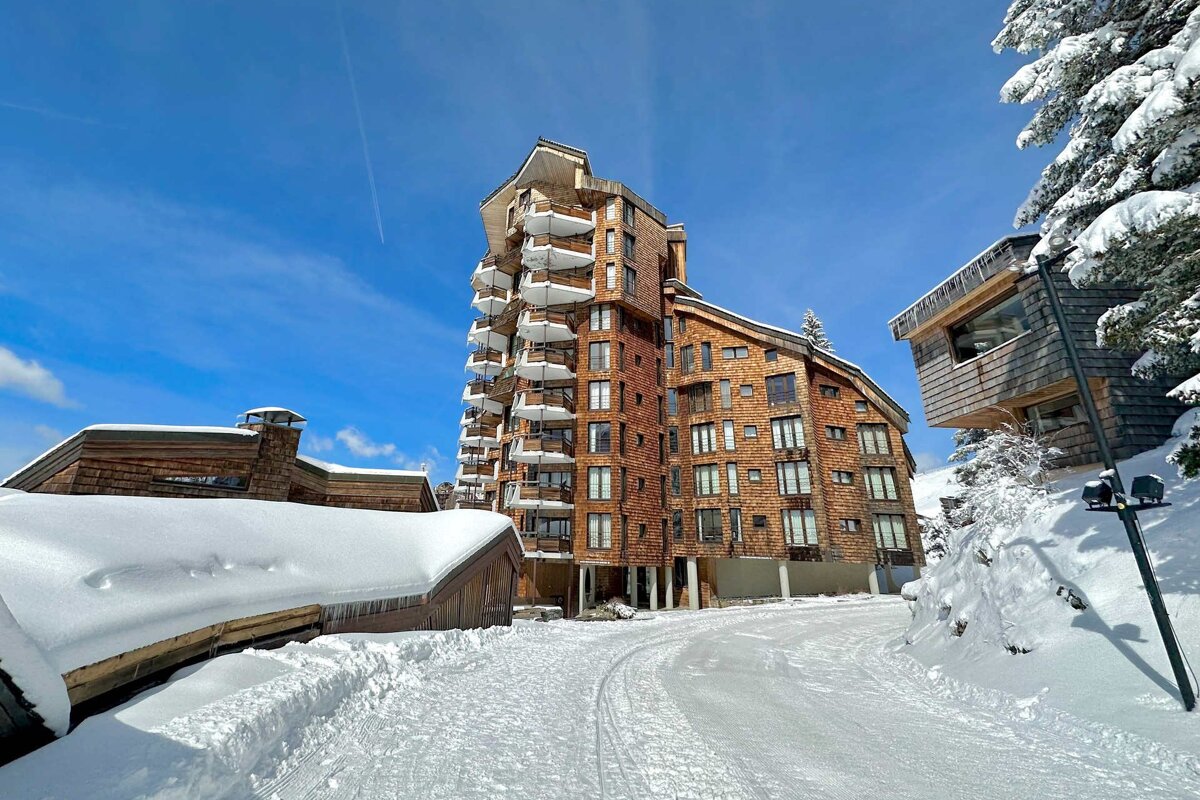 A snowy path leads to striking modern wooden buildings with balconies in a sunny winter ski resort, under a clear blue sky.