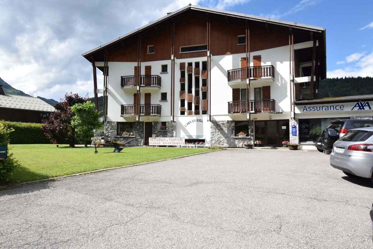 A multi-story chalet-style building with wooden balconies, featuring a green lawn and a paved parking area.