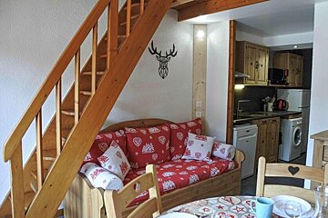 Cozy alpine cabin interior with wooden stairs, a red patterned sofa with pillows, and a small kitchen visible in the background.