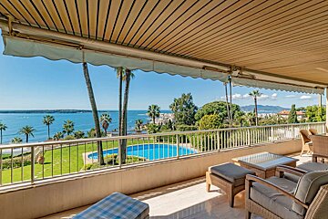A spacious balcony overlooks a vibrant blue sea, palm trees, and a swimming pool in a lush green garden. Outdoor seating is shaded by an awning.