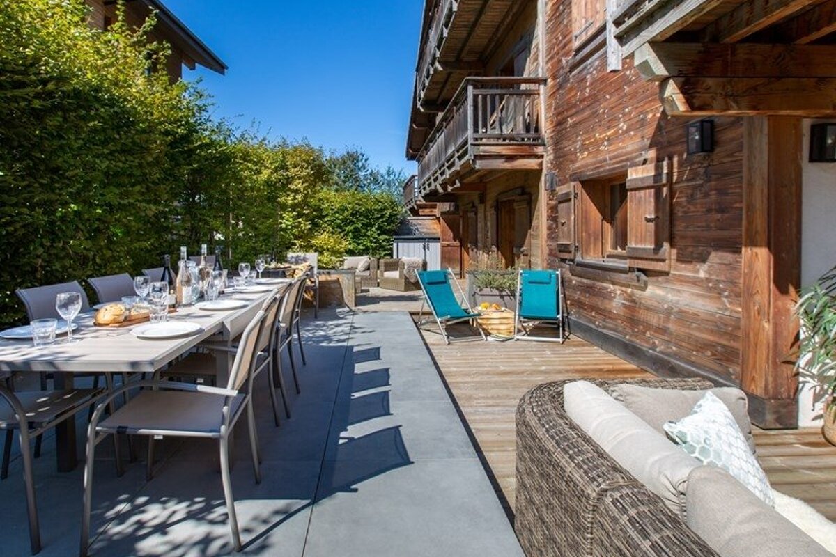 A sunny patio of a wooden chalet featuring a long set dining table, a wooden deck with lounge chairs, and a wicker sofa.