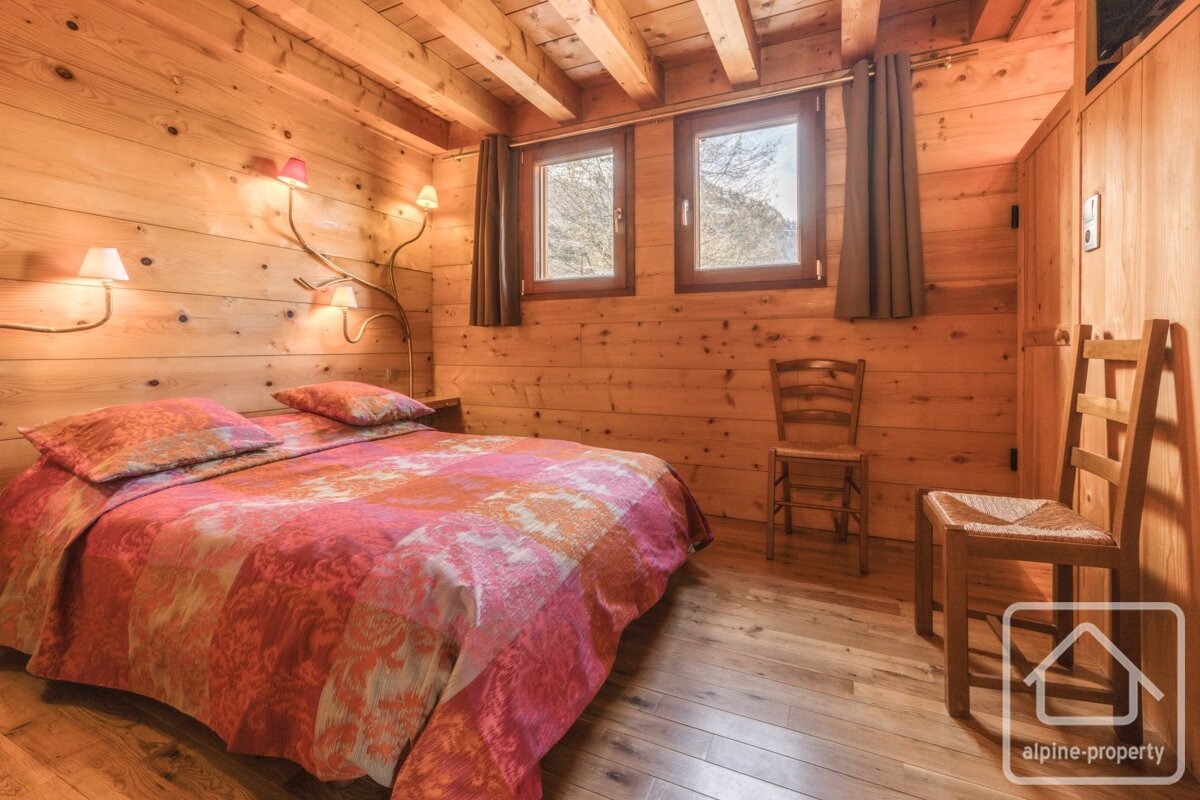 Rustic wooden bedroom with a colorful bed, two windows, unique branch-style lamps, and wooden chairs, exuding cozy chalet charm.