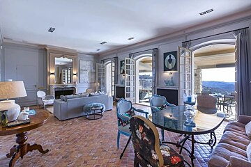A luxurious living and dining room with gray decor, terracotta floors, ornate furniture, and multiple French doors opening to a scenic mountain vista.