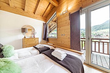 Cozy, wood-paneled bedroom with two single beds, a dresser, and a mirror. A balcony offers scenic views of mountains and houses.