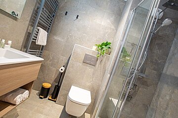 Modern bathroom with grey tiling, featuring a wall-hung toilet, wooden vanity, and glass shower cabin.