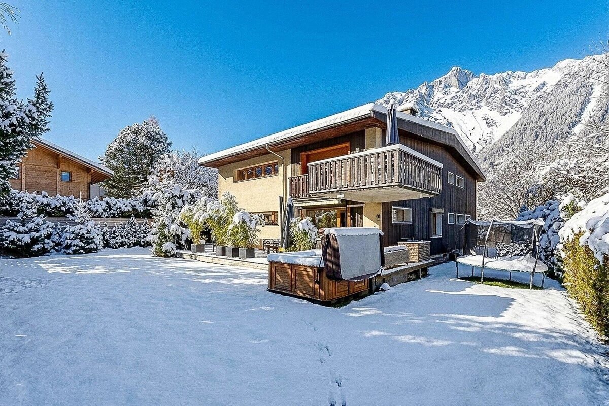 A house with a balcony and hot tub in a snowy yard, surrounded by snow-dusted trees and majestic mountains under a clear blue sky.
