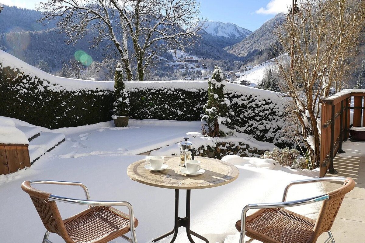A sunny, snow-covered patio with a table, chairs, and coffee, overlooking a beautiful mountain valley in winter.