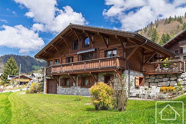 A rustic wooden chalet with stone accents and a balcony, fronted by a green lawn. Mountains are visible under a bright blue, cloudy sky.