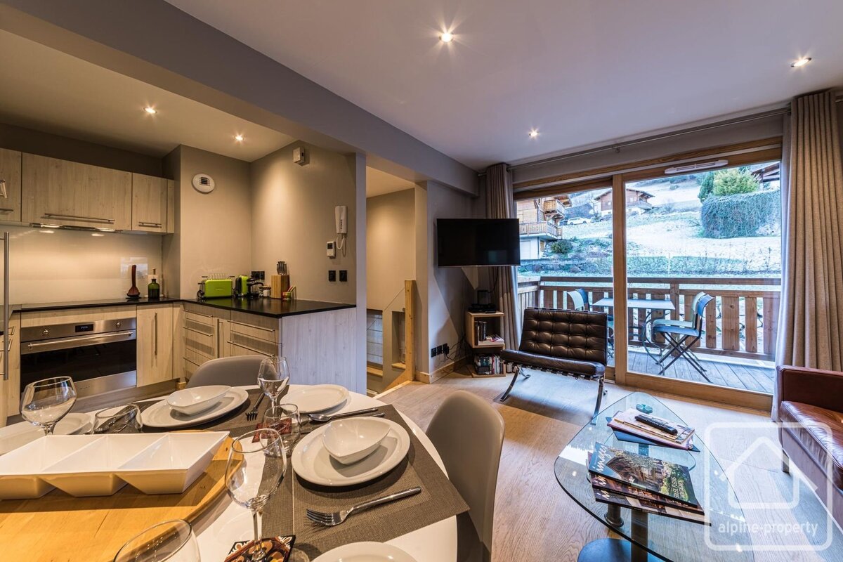 Modern alpine apartment featuring an open-plan kitchen and living area, complete with a dining table and balcony doors showing snowy views.