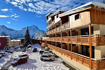 A sunny winter mountain resort with a multi-story building, wooden balconies, snow-covered cars, and majestic peaks under a blue sky.