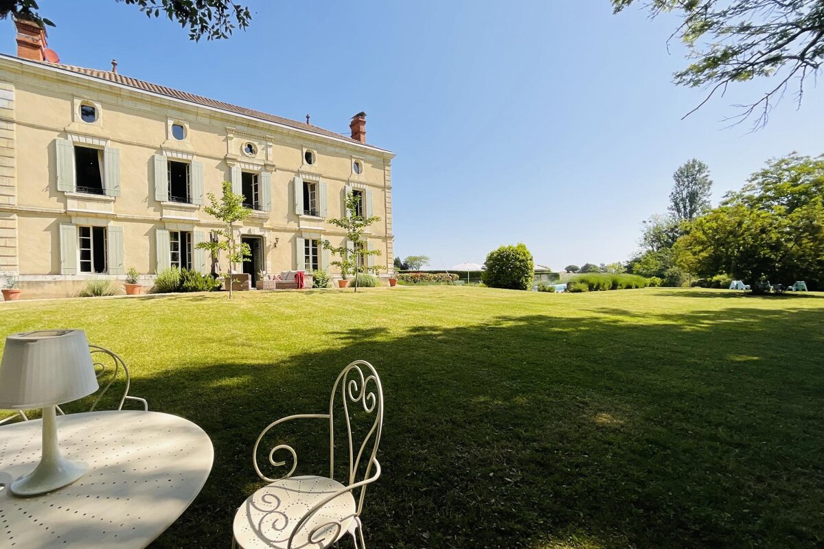 A grand, pale yellow manor house with green shutters stands beside a vast, sunny green lawn. White wrought-iron patio furniture is visible upfront.