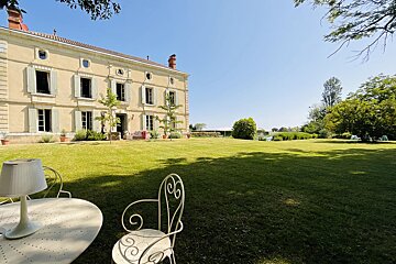 A grand, pale yellow manor house with green shutters stands beside a vast, sunny green lawn. White wrought-iron patio furniture is visible upfront.