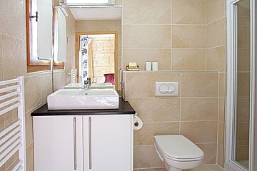 Modern beige tiled bathroom featuring a white vanity sink, a wall-mounted toilet, a large mirror, and a towel warmer.