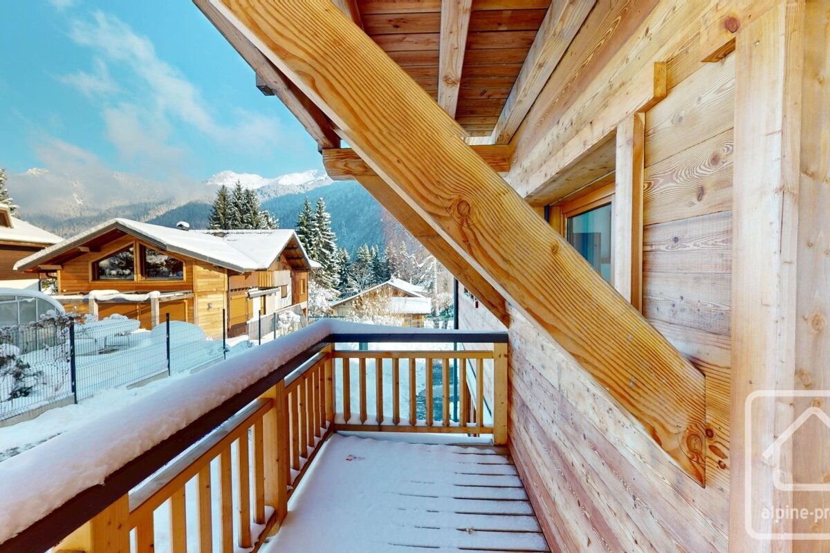 A cozy wooden balcony, dusted with snow, offers a scenic view of a snowy alpine village, evergreen trees, and distant mountains under a bright blue sky.