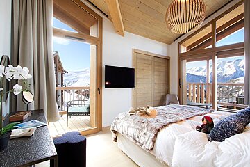 A modern, light-filled bedroom with a cozy bed, wooden ceiling, and expansive windows opening to a balcony with stunning snowy mountain views.