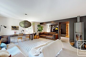 A spacious, modern living room featuring a long wooden desk, two sofas, a fireplace, and a large hanging lamp, with white and dark accent walls.