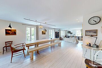 Spacious, bright open-plan room with light wood floors, a long wooden dining table, and a cozy living area in the background, featuring white walls.