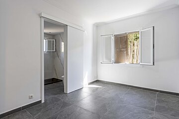 Bright, empty room with white walls, gray tiled floor, and an open window with shutters. A sliding door reveals a bathroom with a toilet.