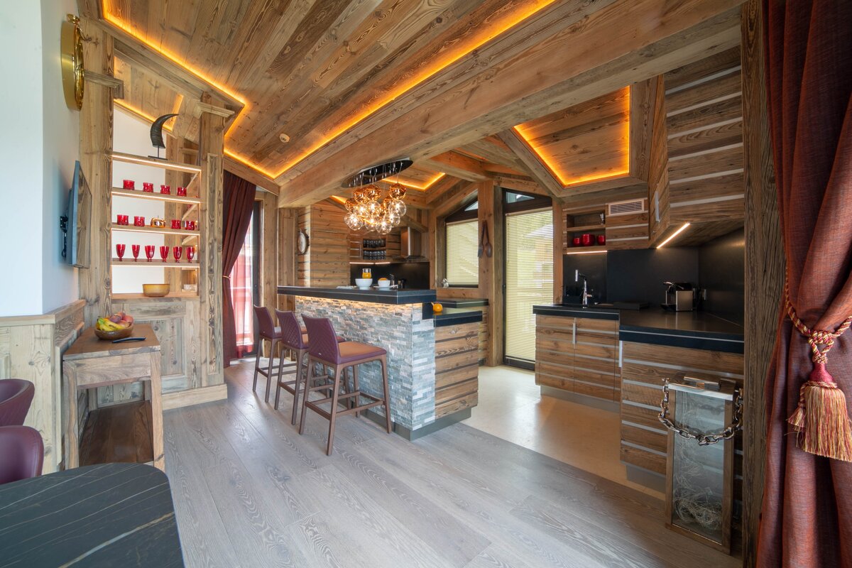 A luxurious, rustic-modern chalet interior with warm wood paneling, illuminated by accent lighting. Features a stylish kitchen/bar with stools and red decor.