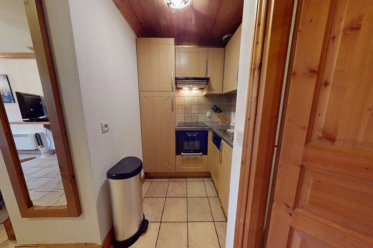 A compact kitchen with light wood cabinets, a dark blue stove, tiled floor, and wood-paneled ceiling. A trash can and mirror are also visible.