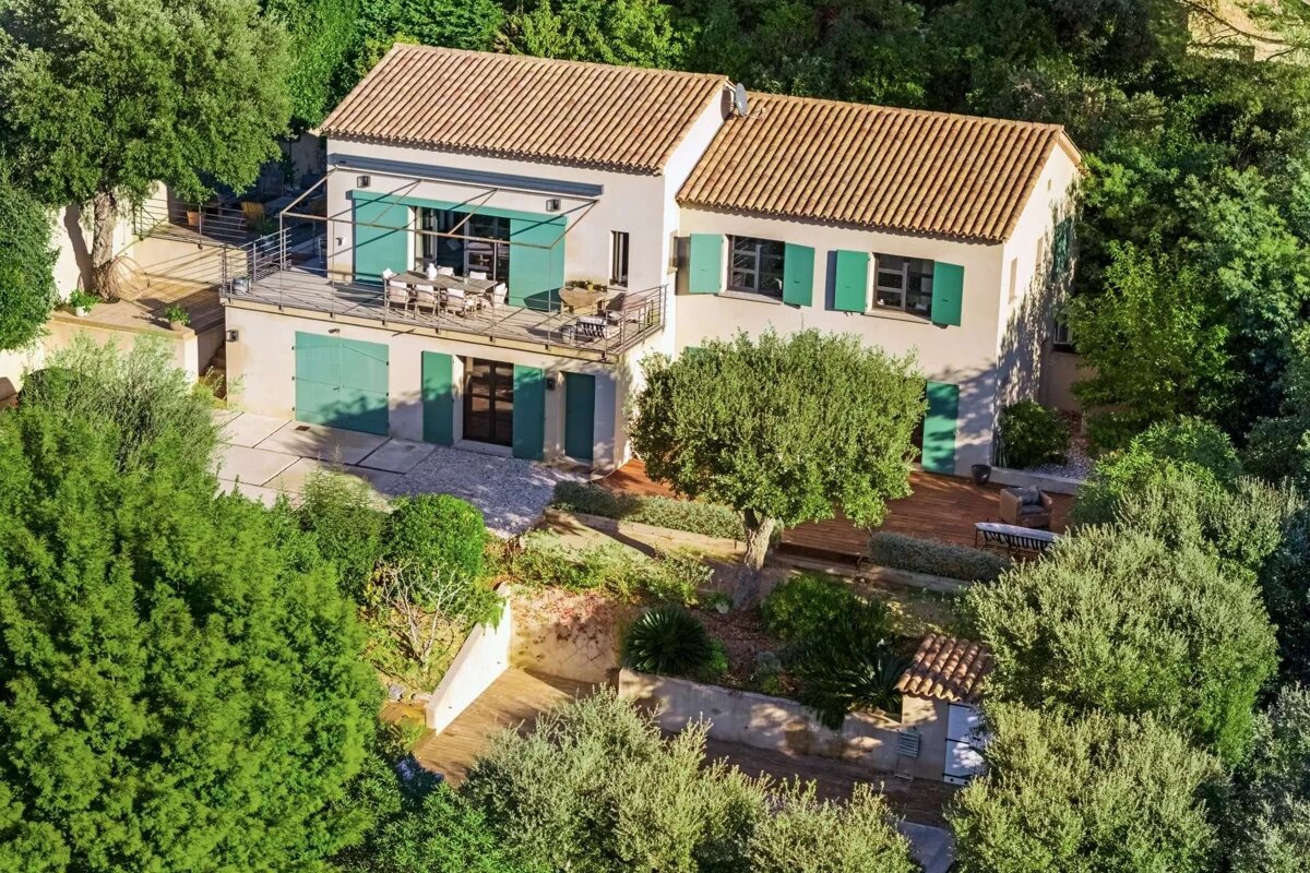 Aerial view of a lovely house with a tiled roof, green shutters, and a balcony, nestled within a lush, terraced garden full of vibrant green trees.