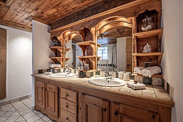 Rustic, warm wooden bathroom with a double vanity, ornate mirrors, open shelves, and tiled surfaces, giving a cozy chalet feel.