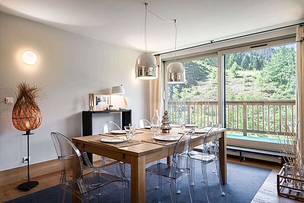 A modern dining room features a wooden table with clear chairs, stylish lighting, and a large window offering a scenic view of a green, forested mountain.