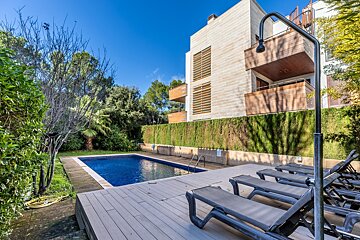 Sunny outdoor pool area with blue water, sun loungers, an outdoor shower, and a modern building with balconies against green hedges.