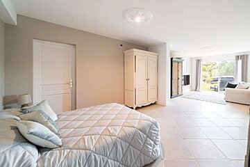 A bright, open-plan bedroom and living area with light-colored walls, tiled floor, a white bed, and a shabby chic wardrobe. A large window overlooks a garden.