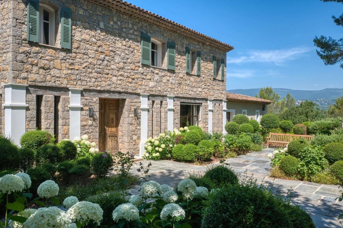 Rustic stone house with green shutters and a lush garden of white hydrangeas, sculpted shrubs, and stone path, offering a distant mountain view.