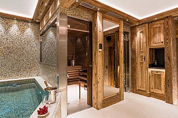 A luxurious indoor spa featuring a tiled plunge pool, champagne, a glass-door sauna, and rustic wood paneling.