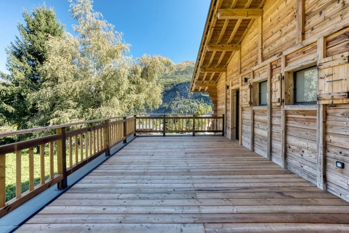 A scenic wooden balcony of an alpine chalet overlooking lush trees and distant mountains under a clear blue sky.
