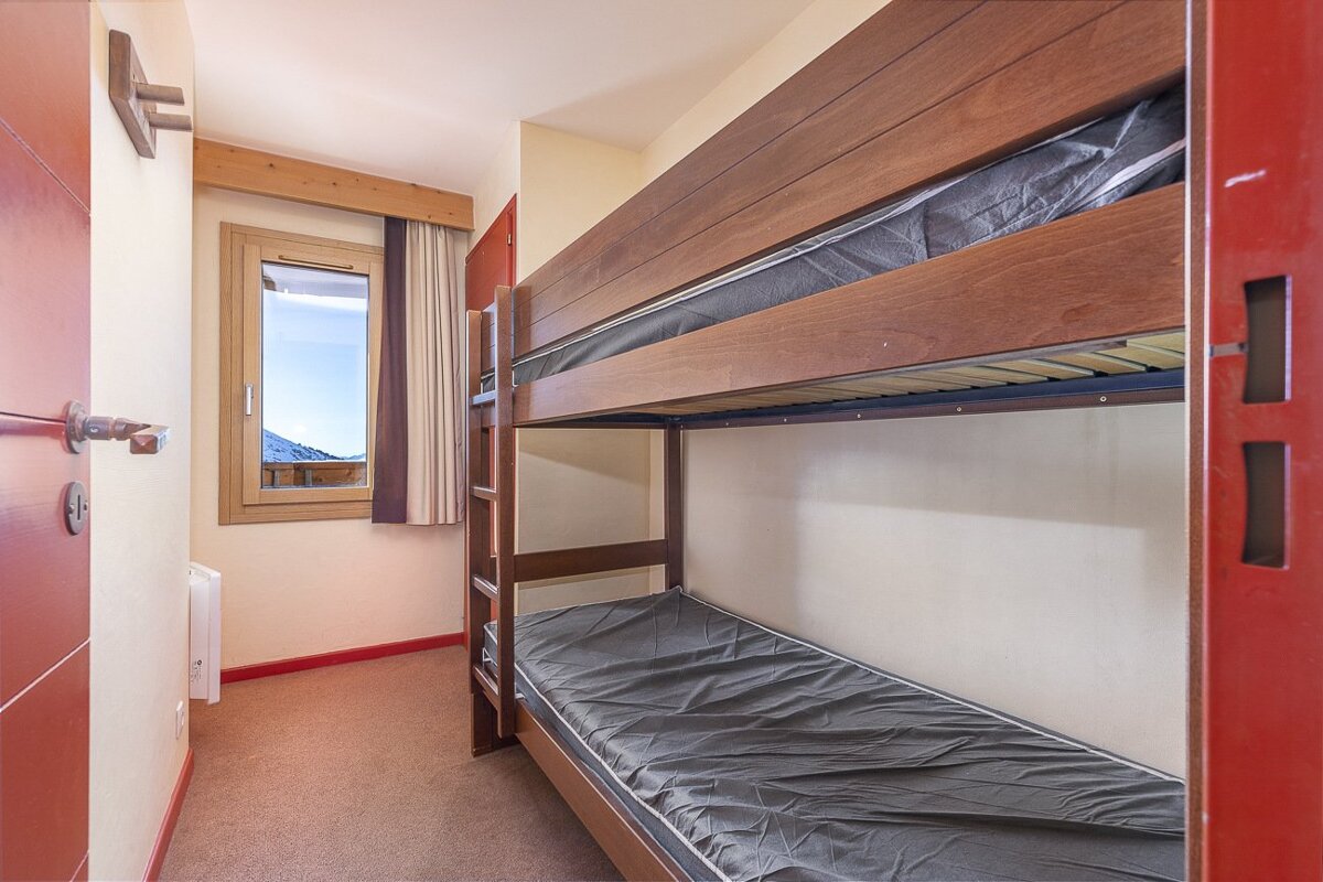 A compact room with wooden bunk beds, a red door, and a window showing snowy mountains. The floor is carpeted.