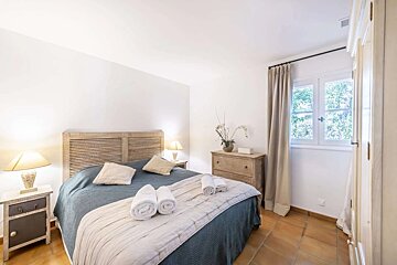 A cozy, bright bedroom with white walls, a bed with a blue spread, wooden headboard, two nightstands, a chest of drawers, and a window with curtains. Terracotta floor.