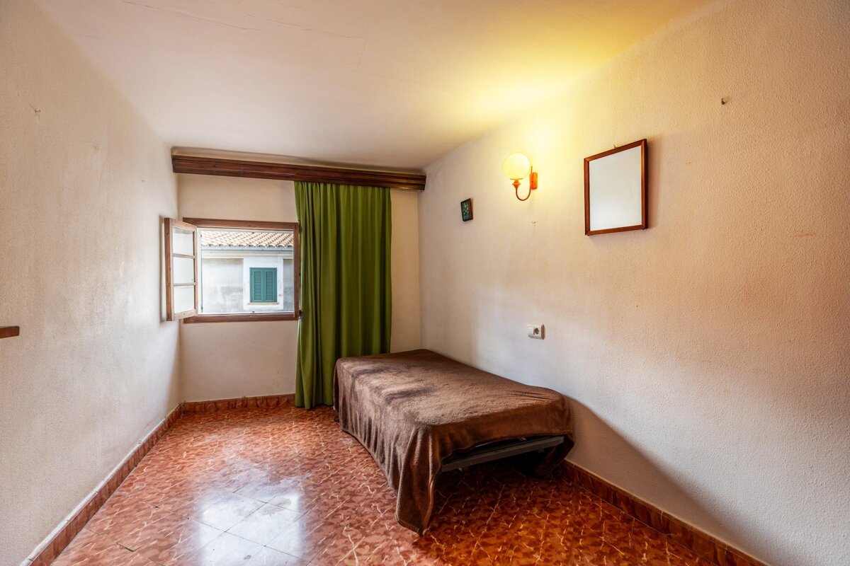 A small, simple room with a single bed, open window with green curtains, and orange tiled floor. Light walls feature a wall lamp and mirror.