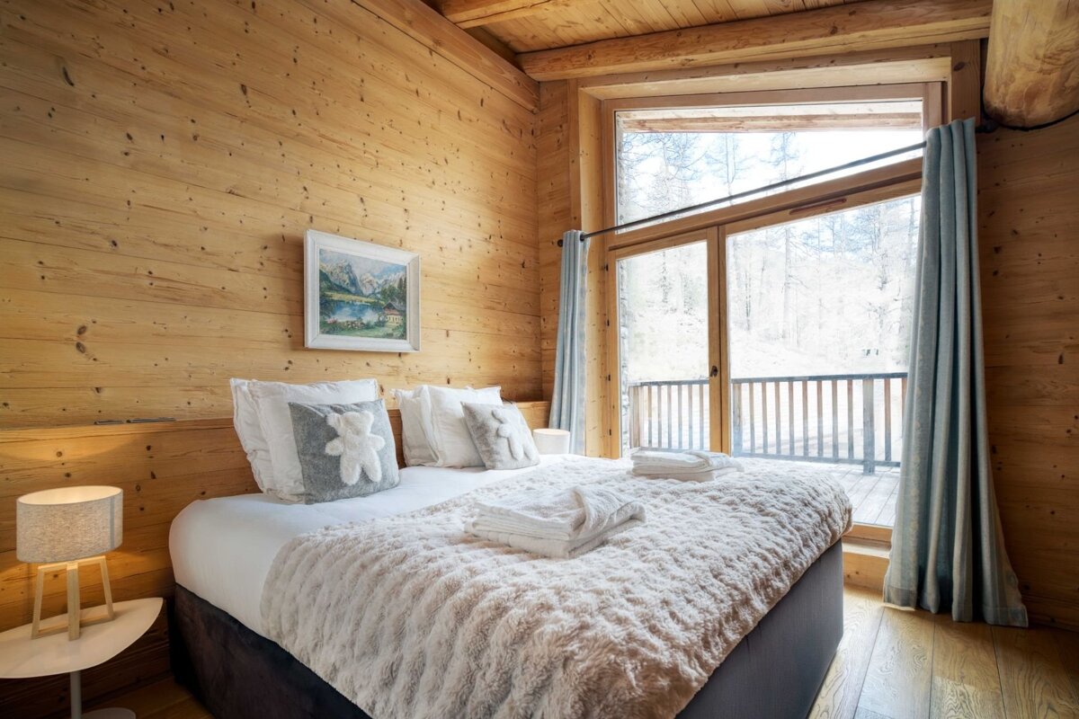 A cozy, wood-paneled bedroom with a large window overlooking a winter forest. The bed features soft white bedding and charming teddy bear pillows.