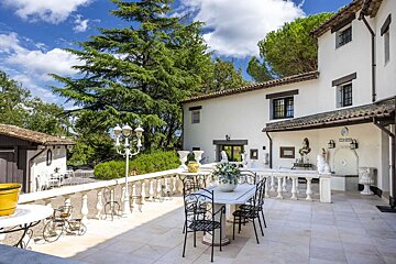 A sunny outdoor patio of a white house features a dining set, elegant balustrade, lush trees, and a blue sky overhead.