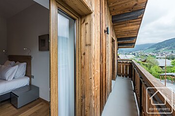 A cozy bedroom connects to a wood-paneled balcony offering panoramic views of a mountainous village landscape under a cloudy sky.