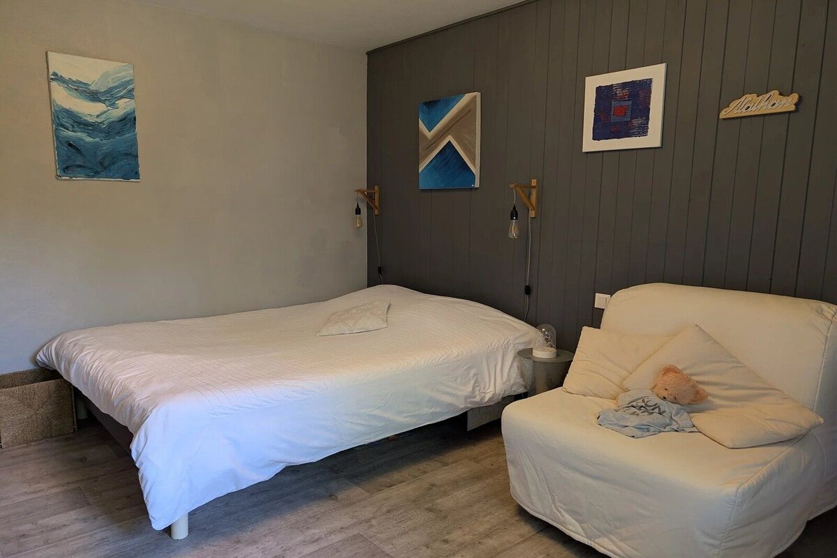 A cozy bedroom featuring a white bed, a grey paneled accent wall with art, and a white lounge chair with a small teddy bear.