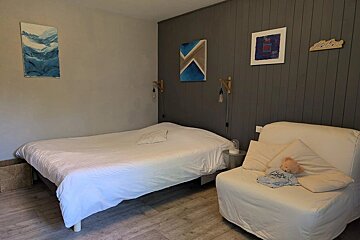 A cozy bedroom featuring a white bed, a grey paneled accent wall with art, and a white lounge chair with a small teddy bear.